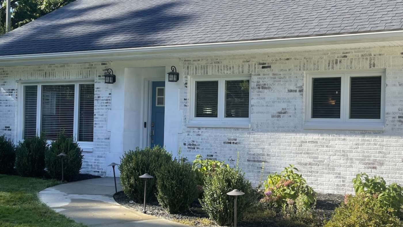 A white-limewashed brick ranch house with a dark shingle roof, a blue front door and a curving front walk, in morning sun.