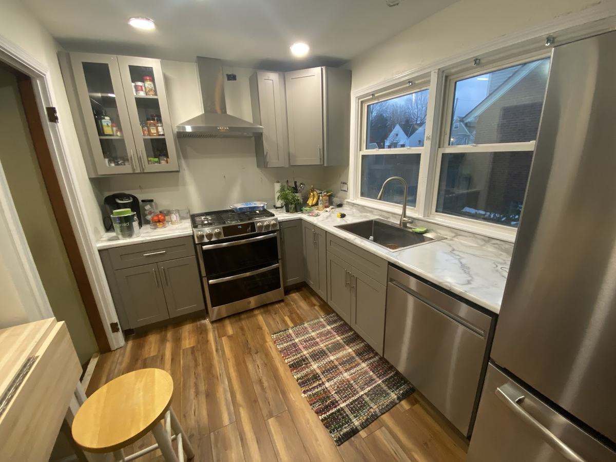 A kitchen with gray shaker cabinets, glass-front uppers, a stainless range under a hood, and a stool on wood-look flooring.