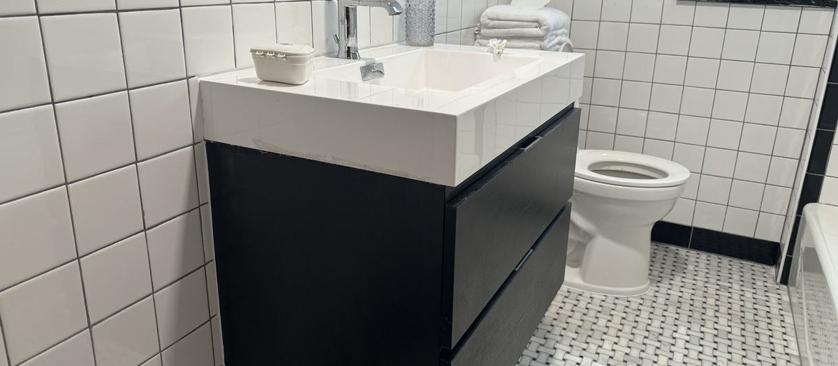 A bathroom with large white wall tile, a black border stripe, a basket-weave tile floor and a black floating vanity with a white sink.
